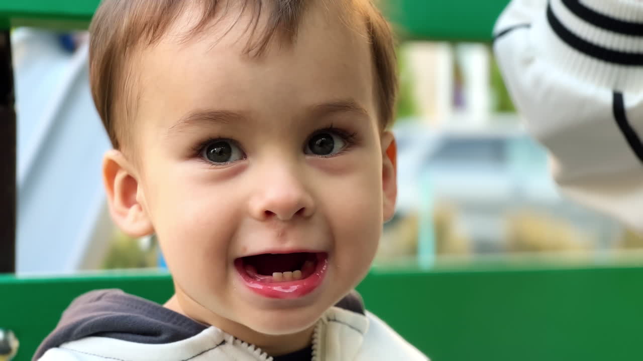 Cute baby opens mouth wide demanding water to drink. Kid sitting on the bench drinks water from bottle. Close up.