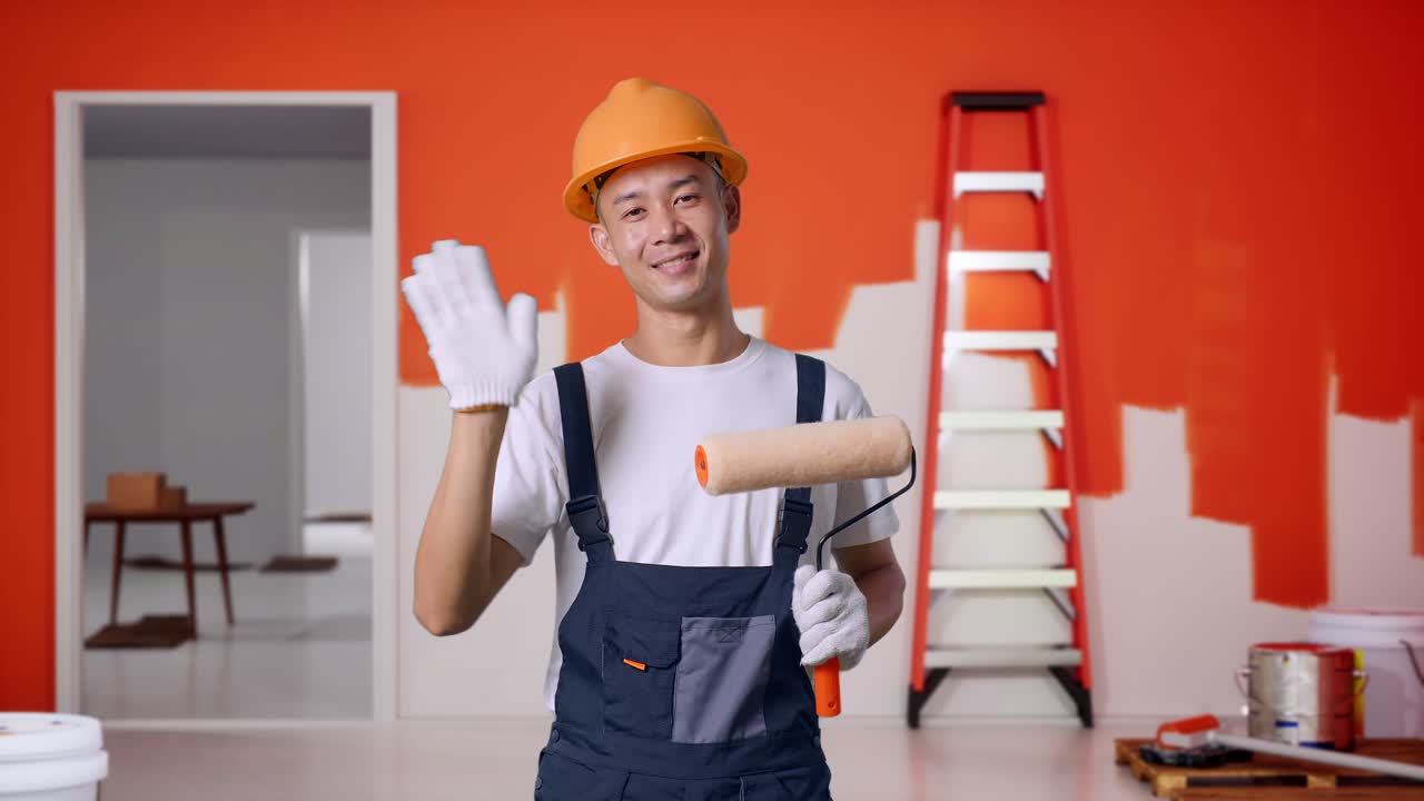 Asian Man Painter Wearing Safety Helmet Smiling, Waving Hand, And Saying Bye While Standing With Painting Wall In Room