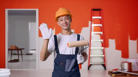 pintor hombre asiático con casco de seguridad sonriendo, agitando la mano, y diciendo adiós mientras está de pie con la pared de pintura en la habitación