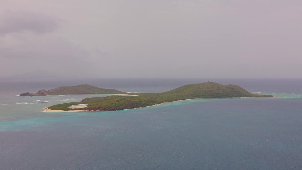 Una ripresa fluida che attraversa l'acqua turchese dell'oceano verso un'isola tropicale lussureggiante e verde sotto un cielo coperto e velato.