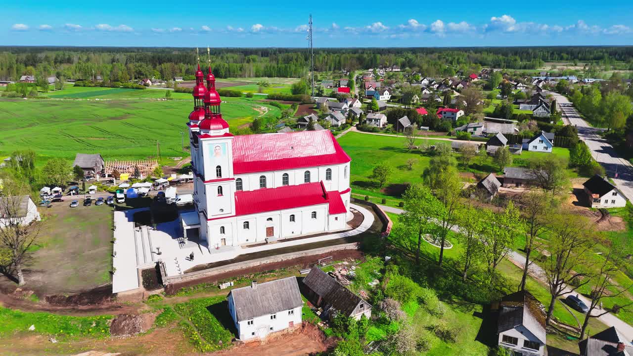 A red-roofed village church with a tall spire stands at the edge of a European rural settlement, framed by farmlands, village homes, and tree-lined roads under a blue sky.