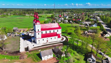 a red-roofed village church with a tall spire stands at the edge of a european rural settlement, framed by farmlands, village homes, and tree-lined roads under a blue sky.