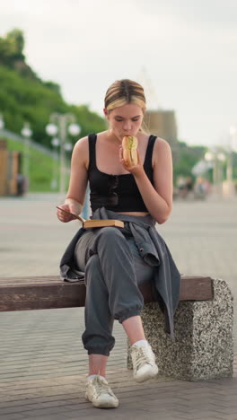 mujer sentada al aire libre con las piernas cruzadas, sosteniendo un libro en una mano mientras come bocadillos con la otra, continúa leyendo, con un fondo borroso con un poste de lámpara y personas caminando