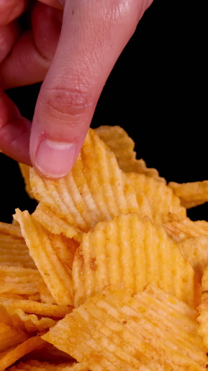 Close-up de mão pegando batata chips ondulada de uma pilha, fundo preto, iluminação de estúdio