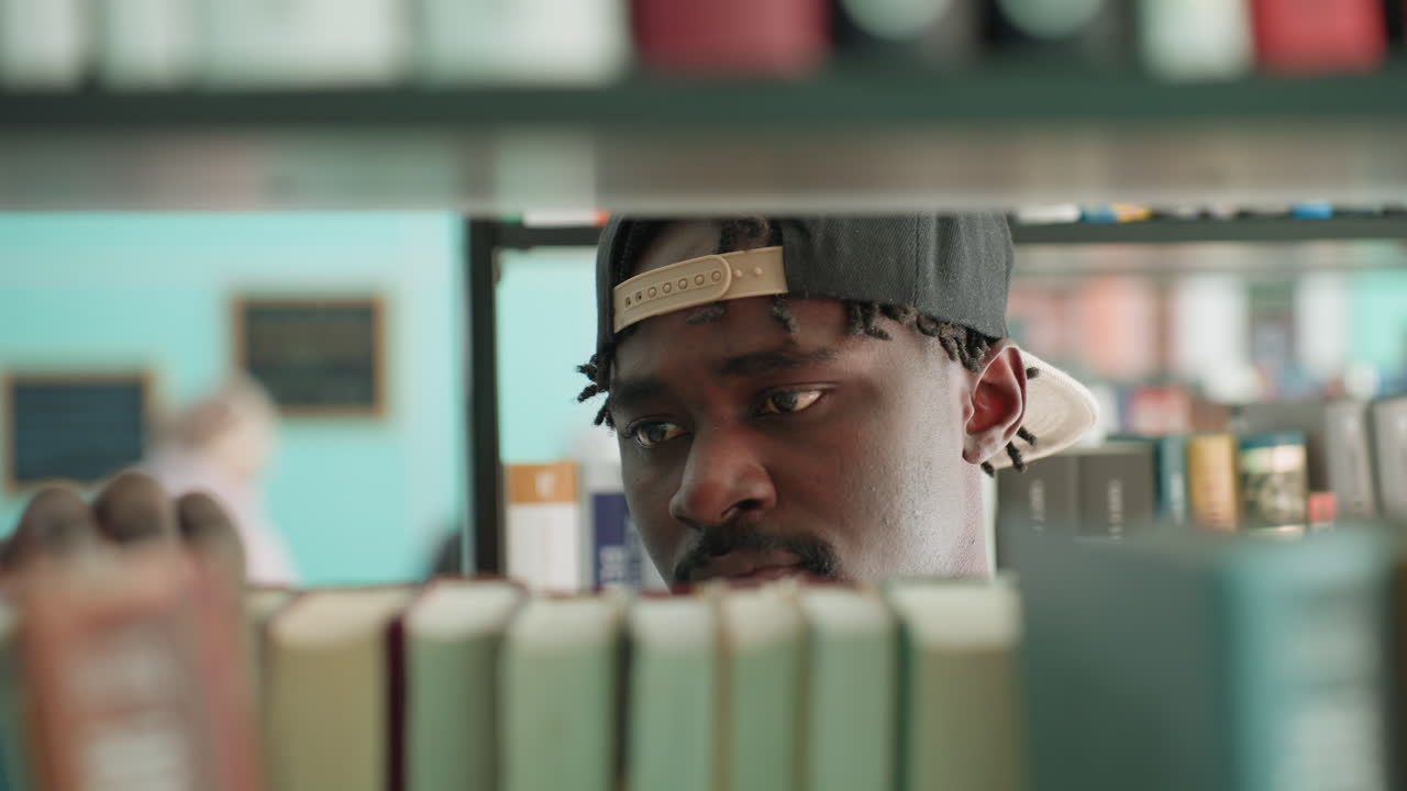 close up in library shows young man in cap scanning row of books, fingers gliding across spines while eyes focus beyond shelf, soft bokeh wall behind