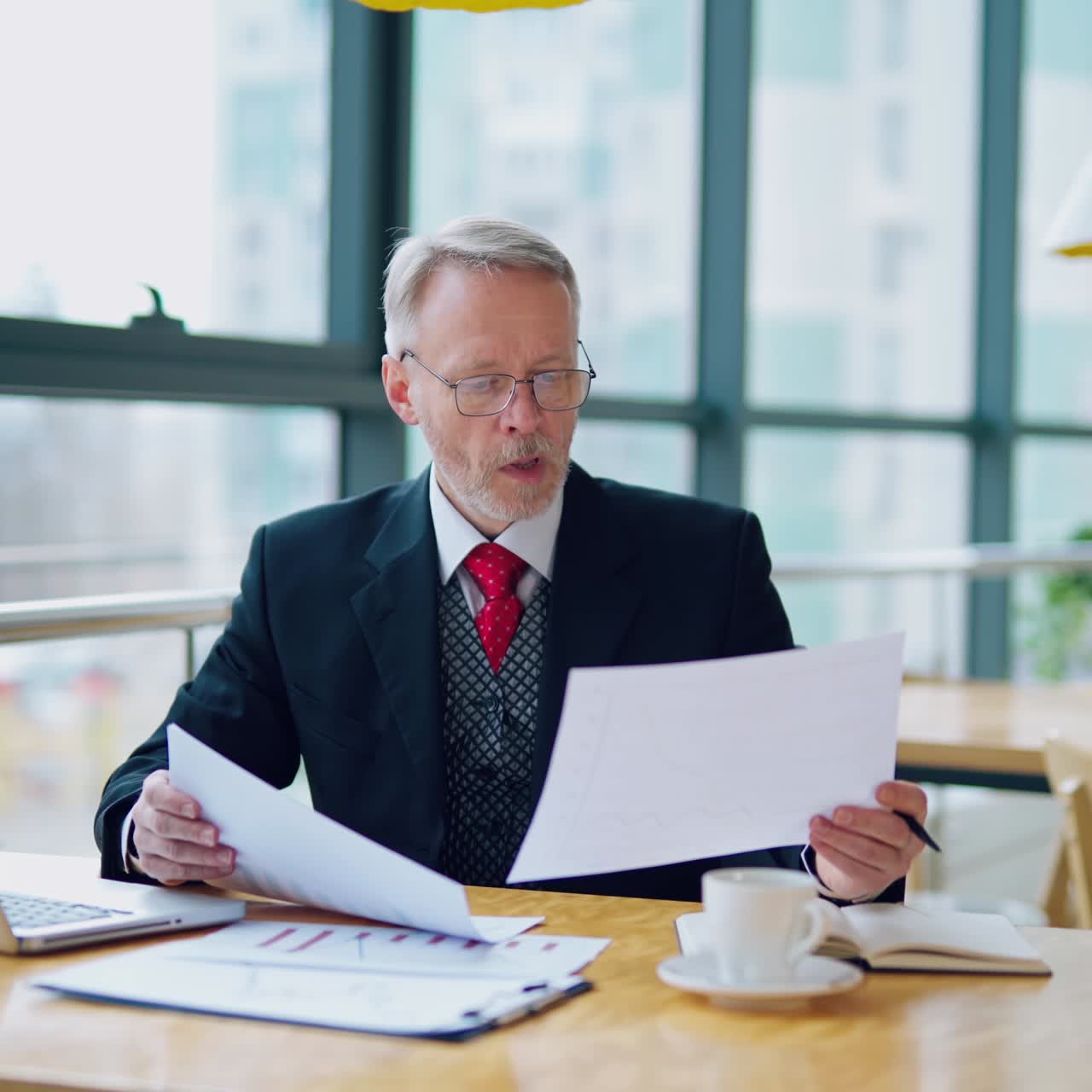 Businessman is reading a printed report or document while sitting at the table, working on paper work with laptop at the table. Man looking at documents. Business video.