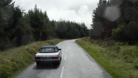 Fly-over-silver-convertible-car-driving-on-road-in-forest-on-cloudy-day.-Starting-to-rain,-raindrops-on-lens.-Ireland