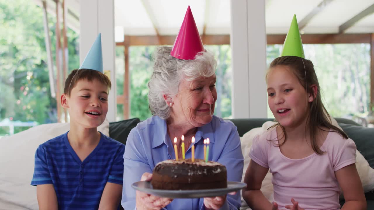 Donna anziana caucasica con cappellino da festa che soffia le candele sulla torta di compleanno mentre i nipoti sono seduti.