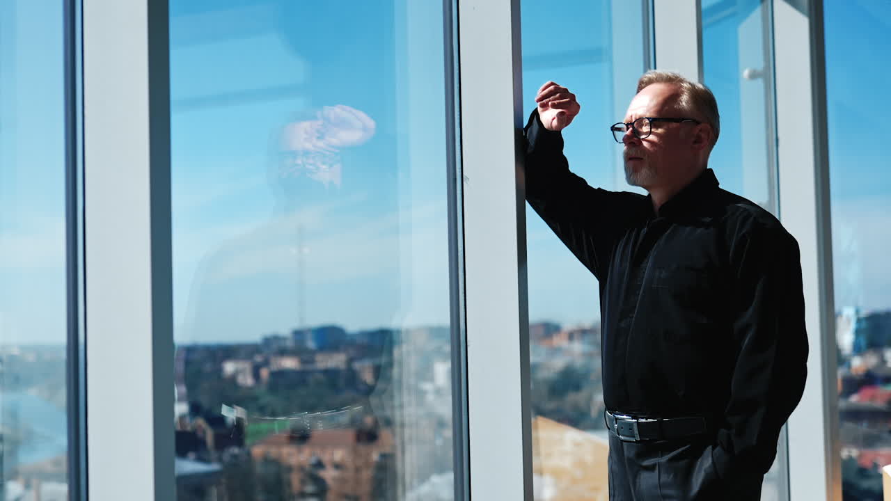 Confident mature Caucasian entrepreneur wearing glasses and black suit. Portrait of a pensive businessman leaning on the window and looking at cityscape.