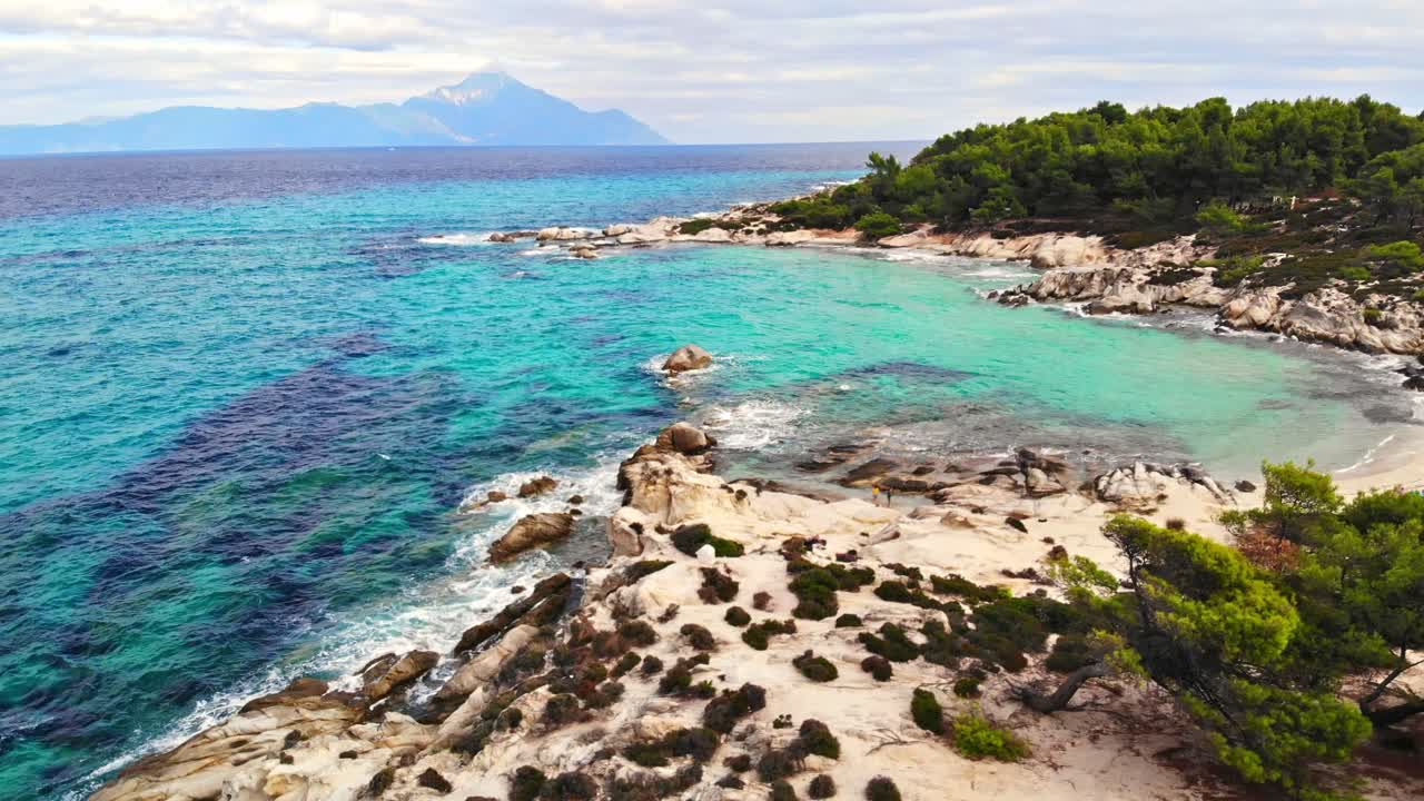 Aegean sea rocky coast with greenery around, blue water and people on the beach, mountain in the distance, Greece
