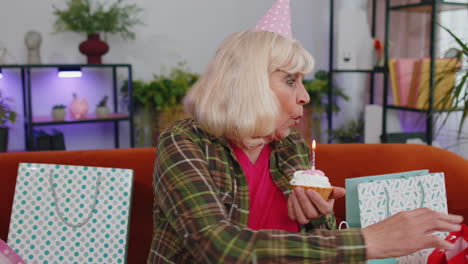 happy senior elderly grandmother woman celebrating birthday party, makes wish blowing burning candle