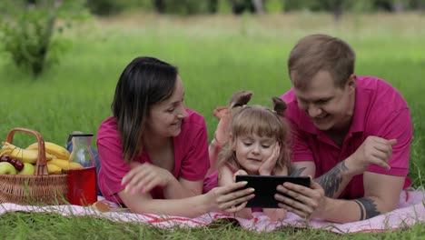 Family-weekend-picnic.-Daughter-child-girl-with-mother-and-father-play-games-on-tablet,-waving-hands