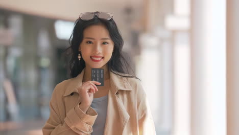 Mujer-Asiática-Feliz,-Joven-Sonriente,-Con-Ropa-Elegante-Y-Moderna,-Usando-Una-Tarjeta-De-Crédito-Y-Bolsas-De-Compras-De-Colores-Brillantes,-Está-Usando-Su-Teléfono-Inteligente-Mientras-Camina-En-El-Centro-Comercial