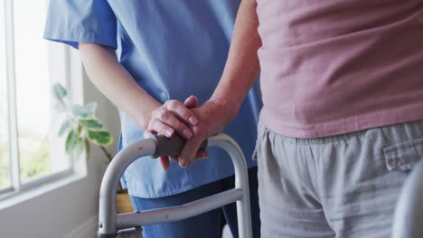 Mid-section-of-female-health-worker-assisting-senior-woman-to-walk-with-walking-frame-at-home