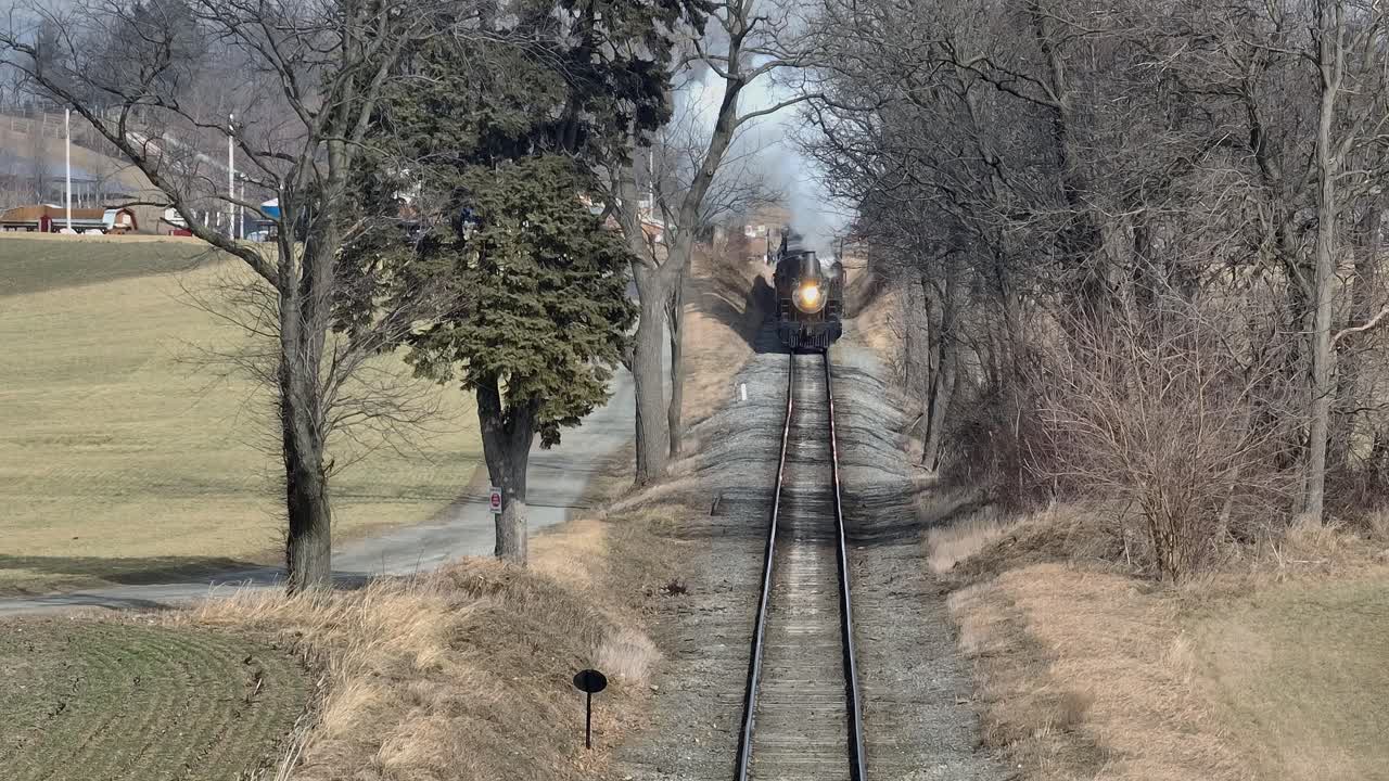 A steam train chugs along the tracks through expansive fields and tall trees in a rural area on a sunny afternoon, creating a picturesque view that evokes nostalgia