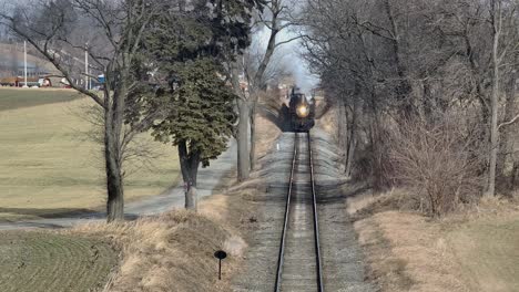 a steam train chugs along the tracks through expansive fields and tall trees in a rural area on a sunny afternoon, creating a picturesque view that evokes nostalgia