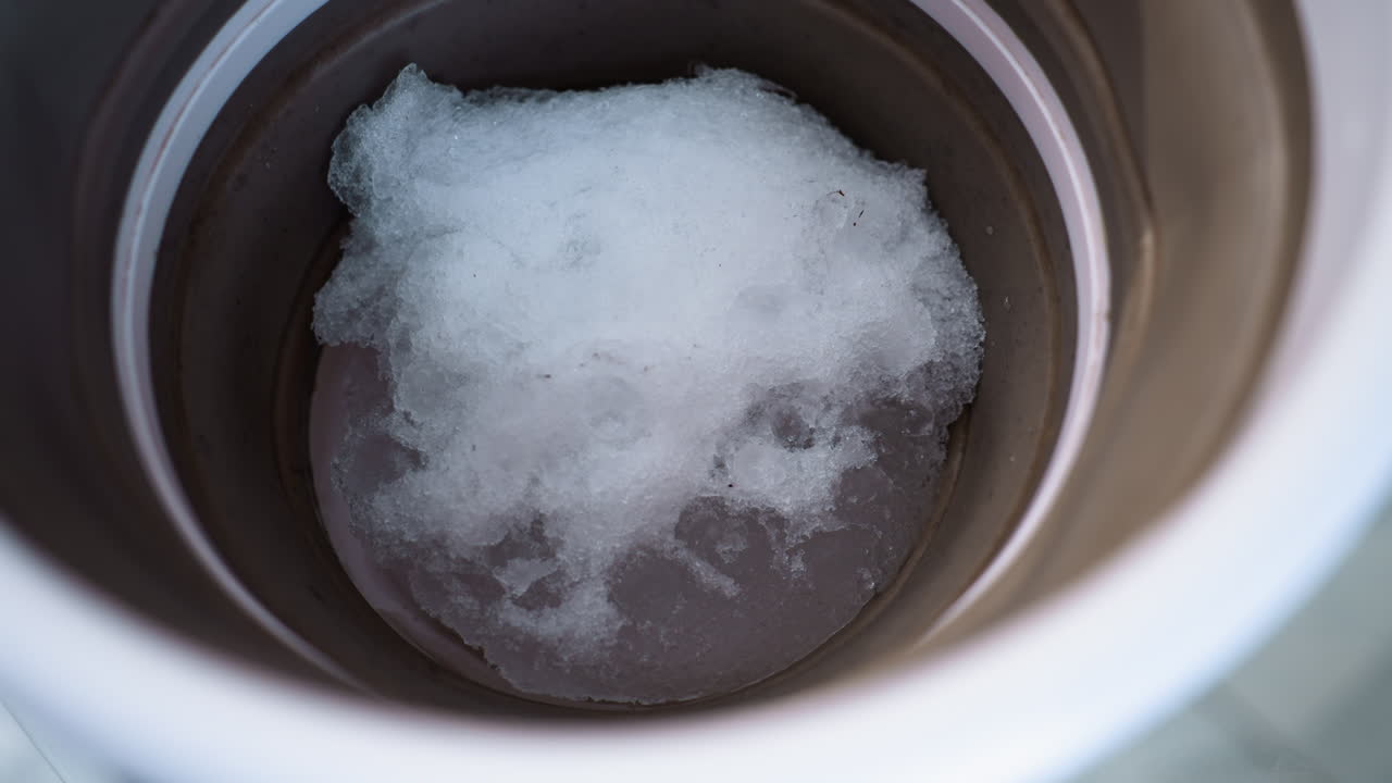High angle close up of brown container on plastic table with melting snow and water droplets, showcasing metallic surface and reflective sheen under soft light creating calm winter mood indoors