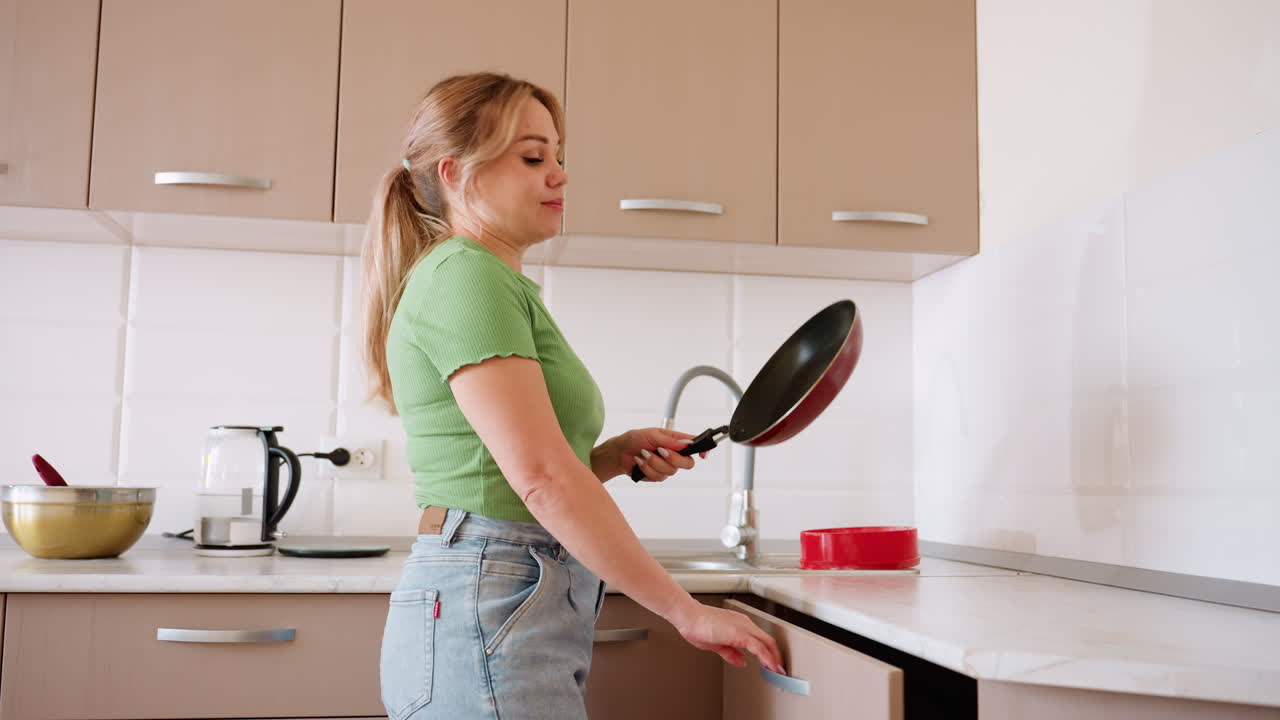 Collaboratrice domestica in maglietta verde e jeans prende una padella dal cassetto della cucina mentre prepara da mangiare, con ciotola in acciaio inossidabile, bollitore e utensili visibili sul piano di lavoro in una moderna cucina luminosa