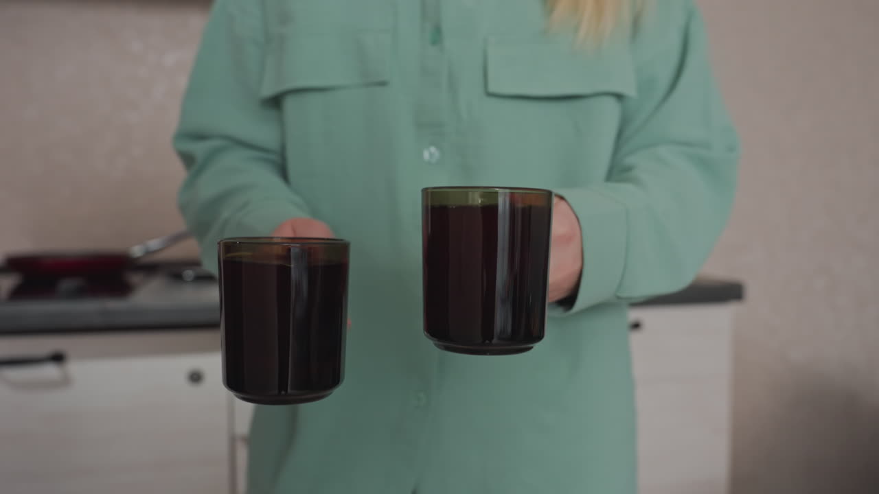 Primo piano di una donna con una manica verde che solleva una tazza di vetro piena di tisana da un tagliere di legno sul bancone della cucina; un bollitore elettrico e barattoli di spezie sono visibili sullo sfondo.