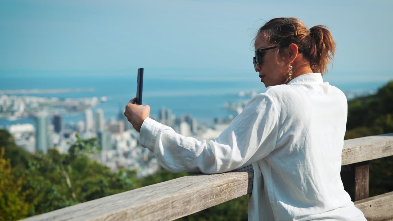 Asian woman with light brown hair, wearing a white shirt and sunglasses, takes a selfie overlooking a vibrant cityscape and ocean