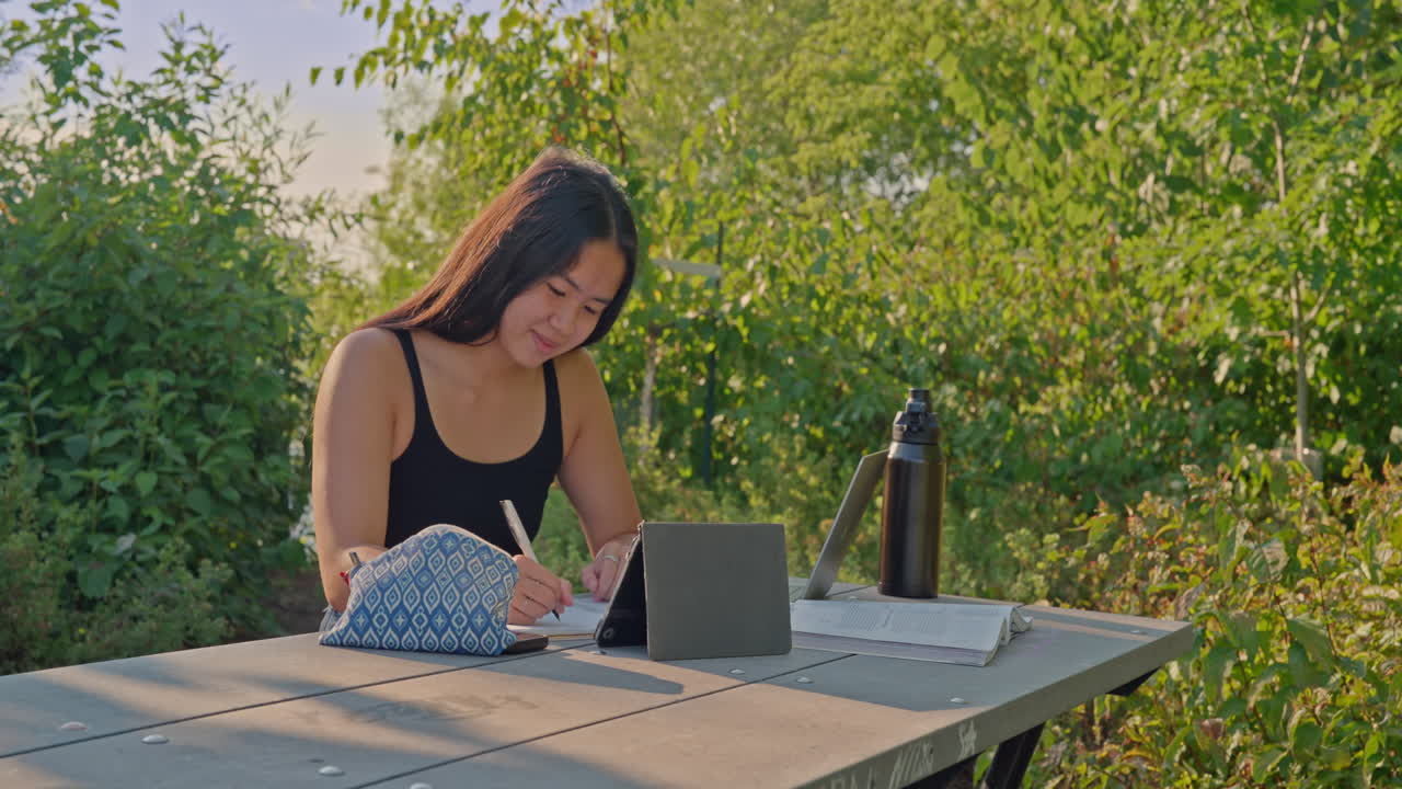 Young Asian student studying outdoors at park table with tablet, notebook and reusable water bottle. Dolly shot moving left, concept of outdoor learning, focus and sustainability