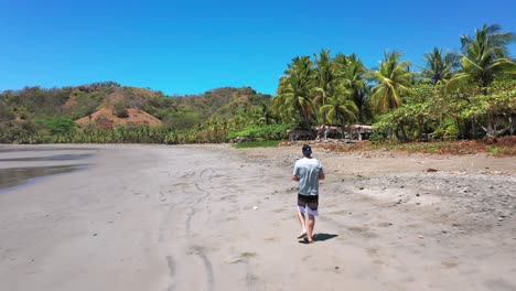 slow motion, follow drone aerial footage of man on wild tropical island, sandy beach in costa rica with palm trees and rainforest landscape, punta islita