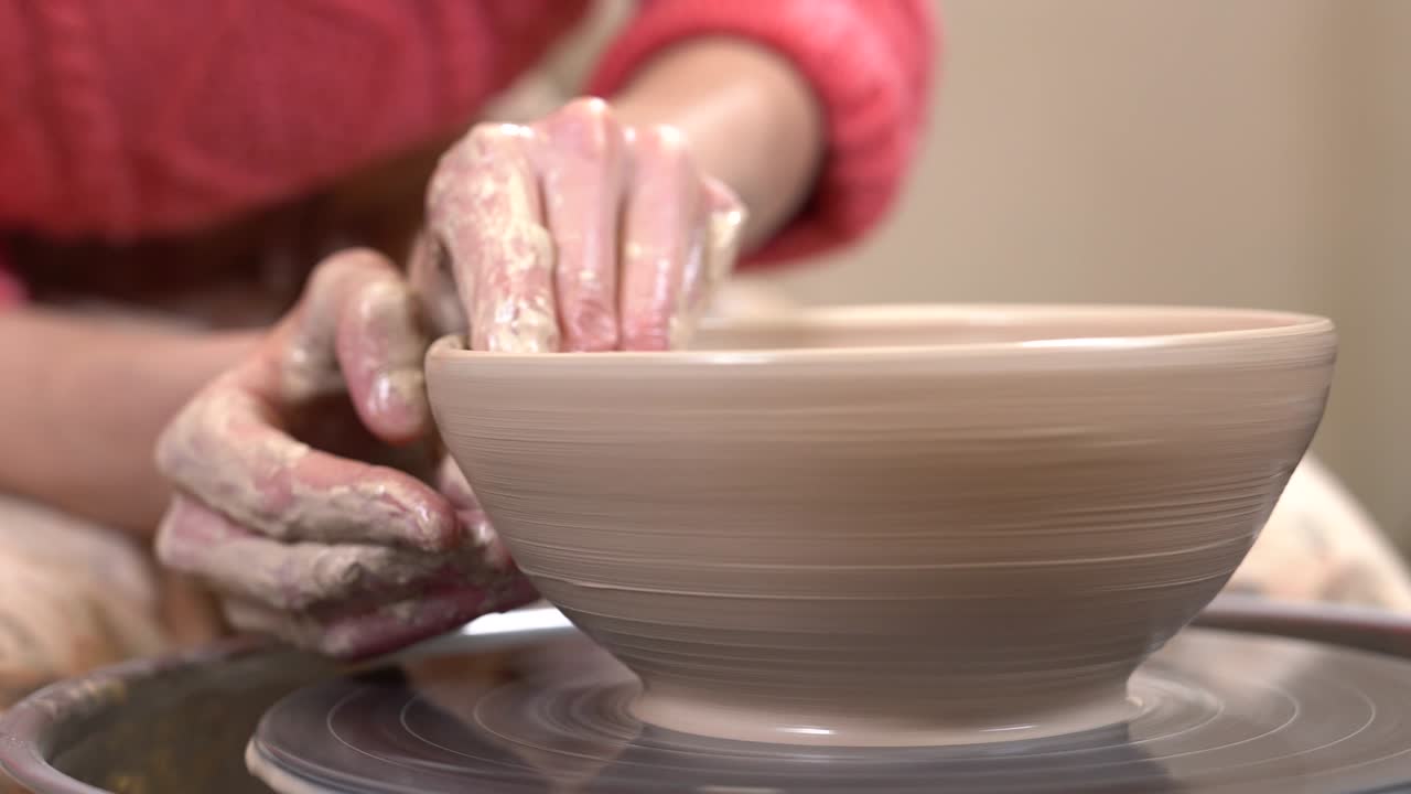 Closeup side view of a muddy hands fine-tuning a clayware shape on the spinning potter's wheel.