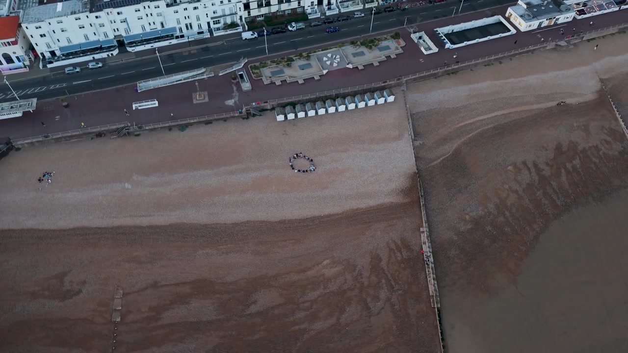Drone gira em vista zenital sobre a praia de Hastings na maré baixa, capturando um grupo de pessoas sentadas em círculo perto do calçadão, Inglaterra.