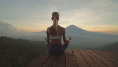 behind fit blond woman sitting in easy lotus pose with silhouette of mount agung