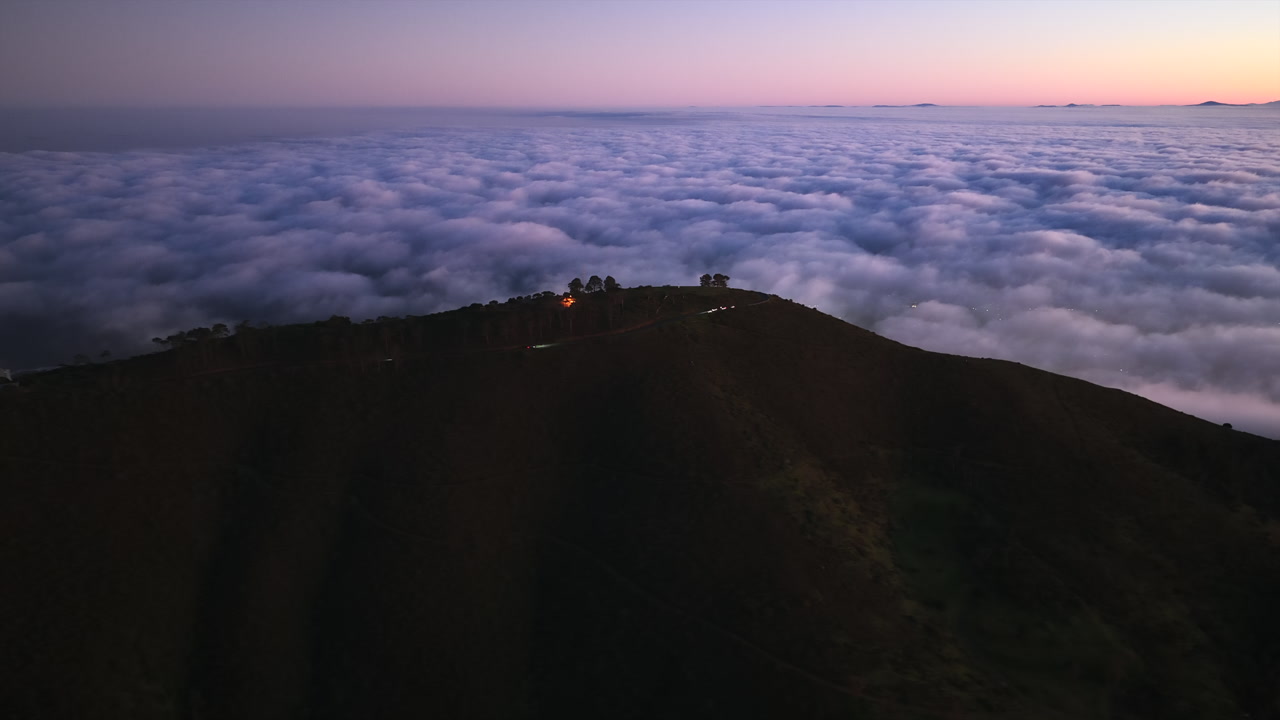 From above Signal Hill, a drone captures sunrise over an endless ocean of clouds, the peak dotted with trees and glowing under morning light.