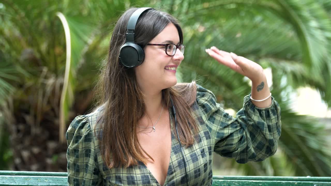 bella chica con auriculares sentados en el banco saludando a alguien y sonriendo en el parque al fondo de una palmera