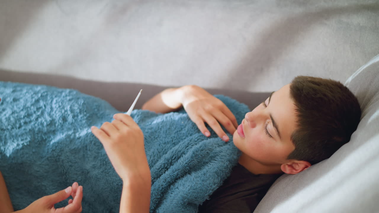 Primer plano de un niño enfermo cogiendo un termómetro para comprobar su temperatura, cubierto con una manta, signo de enfermedad, cuidado en casa, control de salud, descanso y supervisión