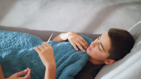 close-up of sick boy collecting thermometer from someone to check his temperature while covered with blanket, sign of illness, at-home care, health check, resting and monitoring