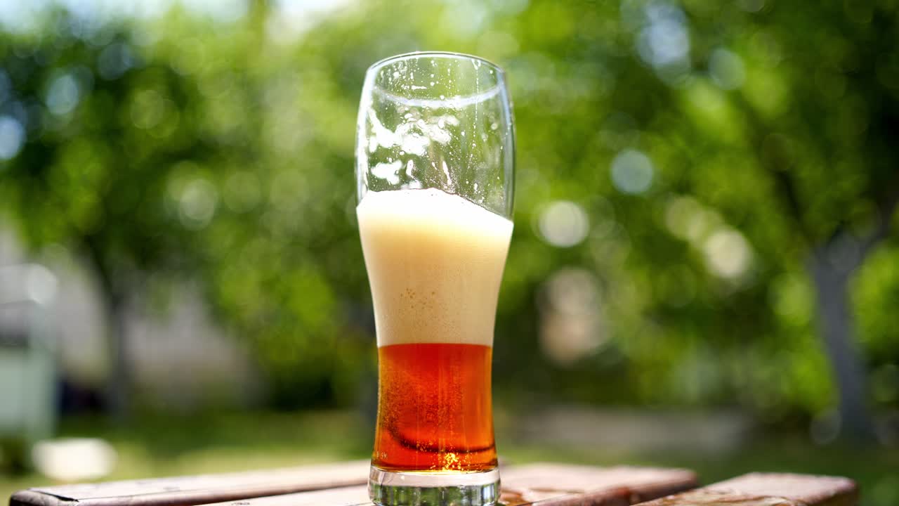 Half glass of cold beer with white foam outdoors. Close-up dark beer with bubbles on blur green background in summer.