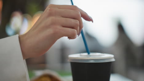 Close-up of hand gently inserting blue straw into takeaway coffee cup lid in vibrant indoor setting with blurred background and glowing bokeh lights, illustrating sustainability and modern lifestyle