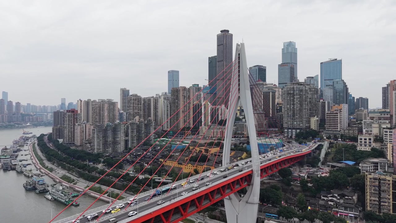 Panorâmica lenta para a esquerda, vista aérea do horizonte de Chongqing a partir da ponte estaiada vermelha sobre o rio, com as densas torres do centro financeiro preenchendo o quadro enquanto a ponte desliza para a direita, tempo nublado, Chongqing, China
