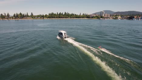 Pontoon-boat-pulling-two-kids-on-a-tube-on-a-lake-with-city-in-background