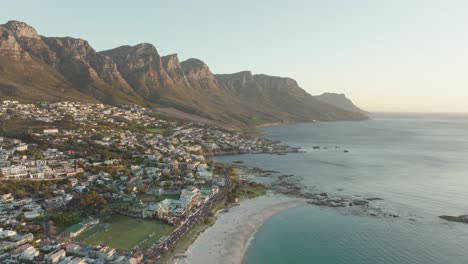 drone flies high above camps bay in cape town south africa - many houses on a hillside - view of table mountain and the endless ocean at sunset