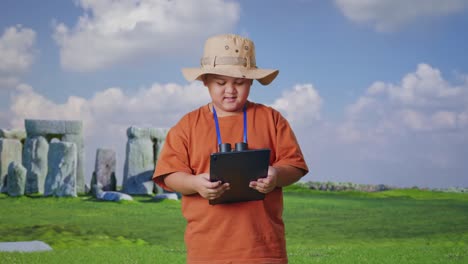 asian boy with a hat and binoculars using a tablet and smiling while traveling in stonehenge. boy researcher examines something, travel tourism adventure concept