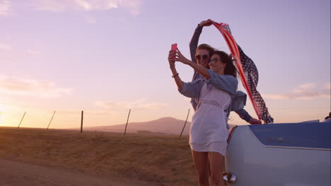 hermosas amigas tomando selfies en un viaje por carretera al atardecer con un coche antiguo