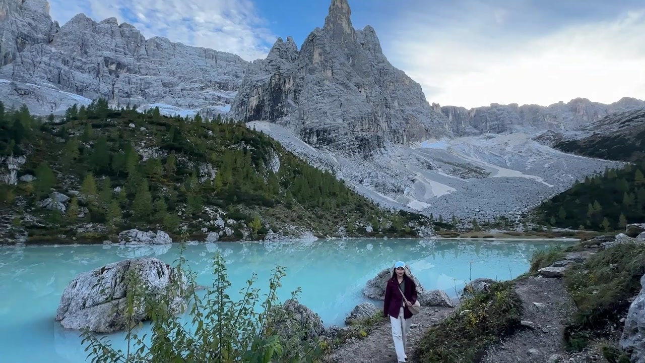 Wide static view of Lago di Sorapis with turquoise water beneath dramatic Dolomite peaks, a woman walking near the shore as soft light and rugged alpine landscape create a serene scene