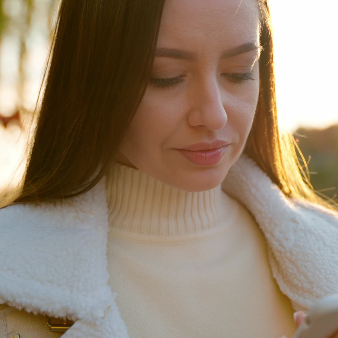 Attractive long-haired brunette looks at her phone typing something. Caucasian woman standing in the rays of autumn sun near the river. Close up