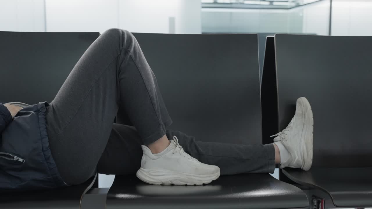 Female passenger in airport terminal lying down tired and exhausted in lounge seats after flight was delayed or cancelled. Workers strike or long wait to board plane at connection gate terminal hall