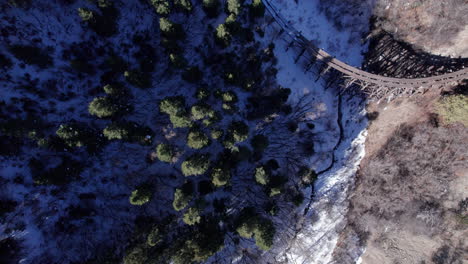 Antena-Arriba-Hacia-Abajo-De-Un-Valle-Cubierto-De-Nieve-En-Un-Bosque-De-Pinos-En-Las-Montañas,-Antiguas-Vías-De-Ferrocarril-De-Vía-Estrecha-En-Primer-Plano