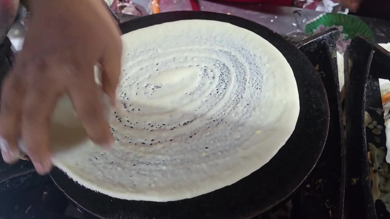 Primo piano di un venditore ambulante che stende la pastella per dosa su una piastra calda, creando una base sottile e croccante durante la preparazione di cibo tradizionale dell'India del Sud in una bancarella affollata
