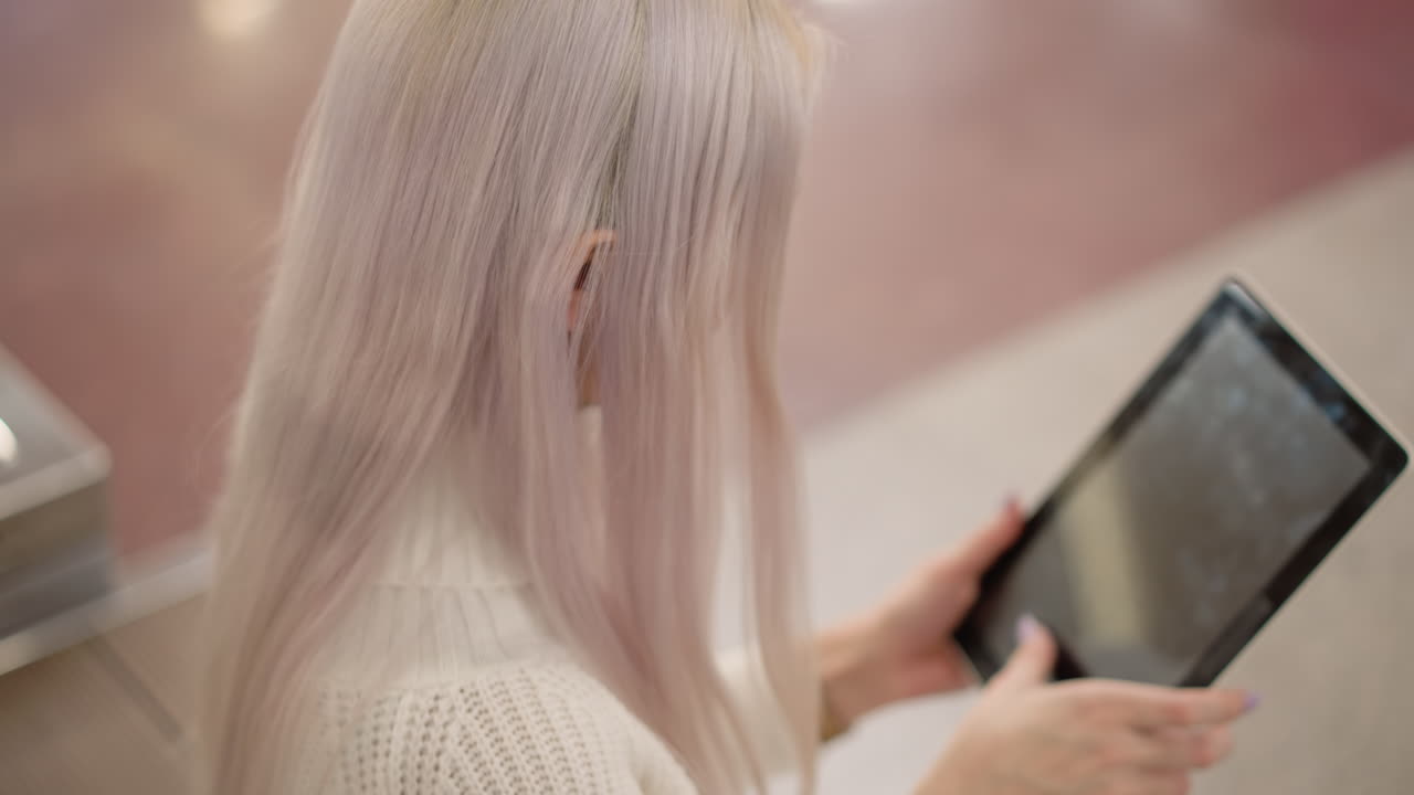 back view of browsing woman on bench using manicured nails to tap tablet screen in spacious mall hall, blurred polished floor elegant manicure highlighting digital interaction with modern device