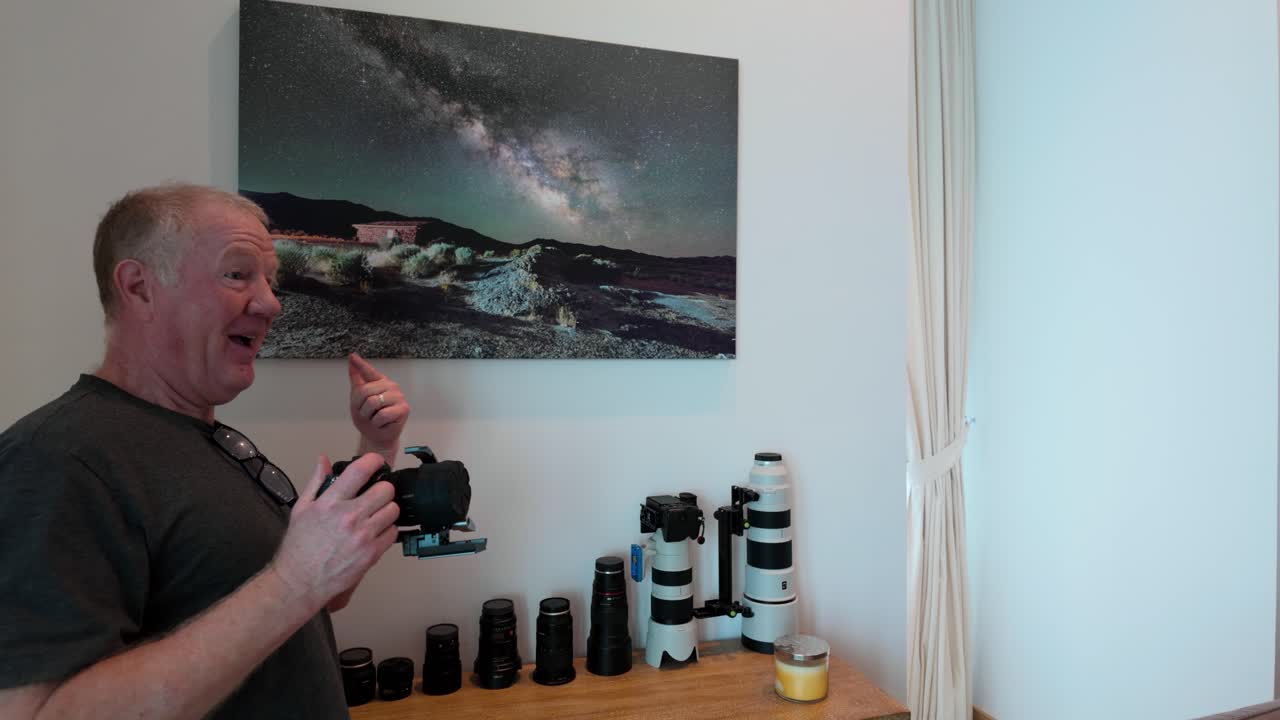 Ældre fotograf beundrer sit Mælkevejsprint og poserer med det. Kameraer og objektiver fremvises på et træbord. Klart, naturligt lys fremhæver den intime atmosfære og kunstneriske stemning.