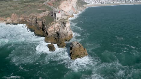 Atemberaubende-Meereswellen-Plätschern-An-Der-Klippenküste-Und-Der-Festung-Des-Heiligen-Erzengels-Michael-In-Nazare,-Leiria,-Portugal