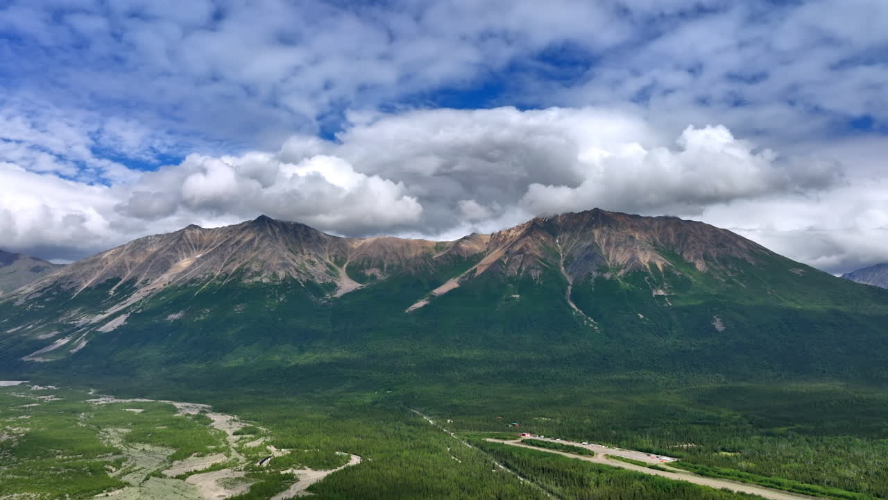 Flight over the vast valley surrounded by the rocks. White fluffy cloudscape accumulated above the mountain tops. Alaska, USA