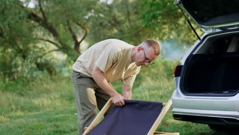 man in glasses having troubles with unfolding the chair. finally man succeeds and sits down in a chair. nature backdrop.